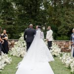 Bride walks down aisle escorted by father toward groom during outdoor ceremony lined with white floral arrangements