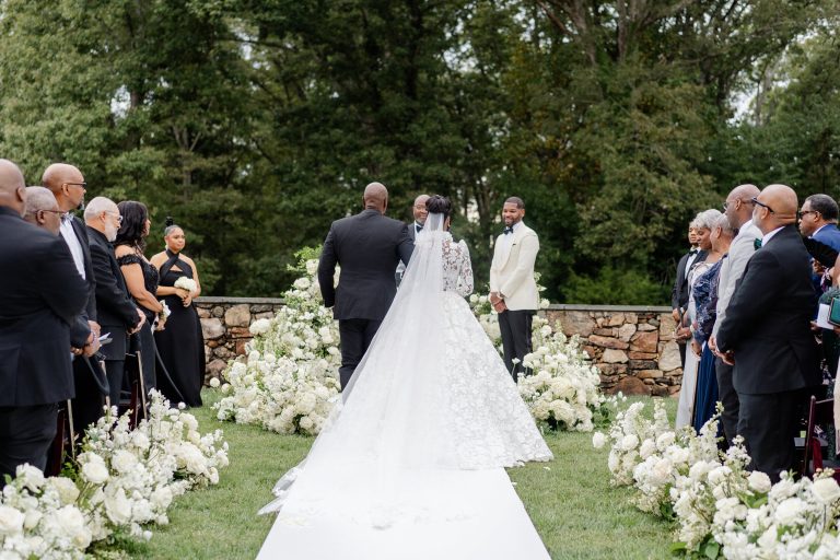 Bride walks down aisle escorted by father toward groom during outdoor ceremony lined with white floral arrangements