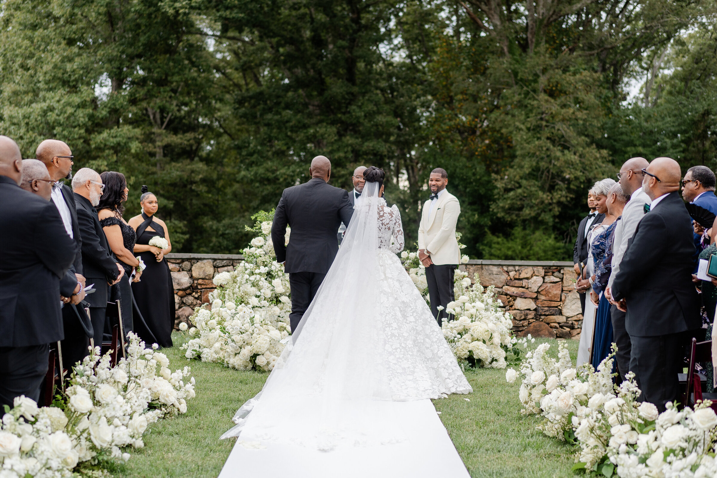 Bride walks down aisle escorted by father toward groom during outdoor ceremony lined with white floral arrangements