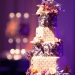 Five-tier wedding cake with quilted fondant, purple scrollwork piping, and cascading peach and purple flowers
