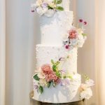 Four-tier white wedding cake with watercolor effect and fresh pink, white, and lavender flowers on gold stand