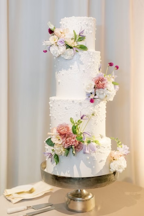 Four-tier white wedding cake with watercolor effect and fresh pink, white, and lavender flowers on gold stand
