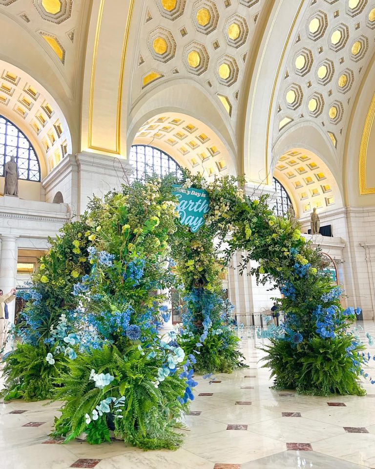 Large floral Earth Day arch with blue hydrangeas and ferns in ornate building hall with coffered ceiling