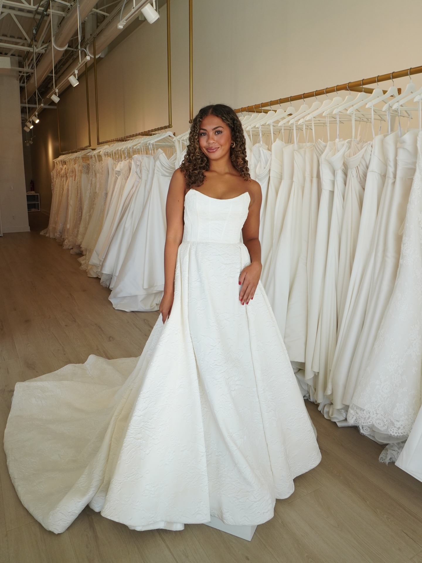 Bride trying on strapless ballgown wedding dress with textured fabric and dramatic train in bridal salon