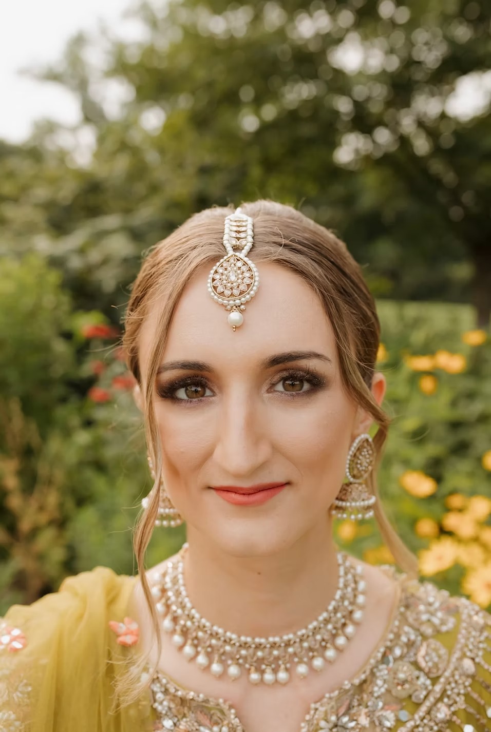 Bride in yellow and gold embroidered South Asian wedding attire with pearl tikka and statement jewelry in garden setting