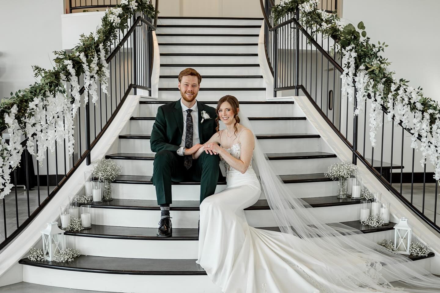 Newlyweds on decorated staircase with cascading white florals, candles, and bride's flowing veil