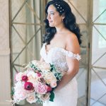 Bride with cascading dark waves and pearl hair clips in lace off-shoulder gown holding romantic rose and peony bouquet