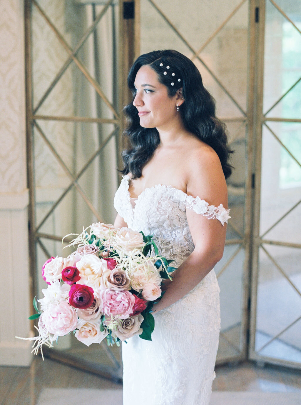 Bride with cascading dark waves and pearl hair clips in lace off-shoulder gown holding romantic rose and peony bouquet