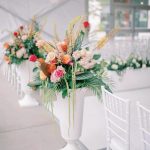 Colorful wedding ceremony arrangement with roses, pincushion proteas, and palm fronds in tall white urn beside chiavari chairs