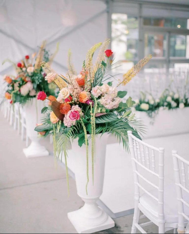 Colorful wedding ceremony arrangement with roses, pincushion proteas, and palm fronds in tall white urn beside chiavari chairs