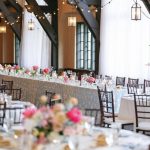 Wedding reception venue with exposed beams, string lights, and pink floral centerpieces on white-clothed tables