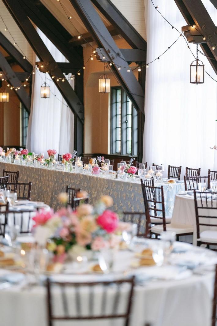 Wedding reception venue with exposed beams, string lights, and pink floral centerpieces on white-clothed tables