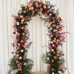 Vibrant wedding ceremony arch adorned with coral, pink, and yellow roses accented by dark foliage and tropical palm fronds