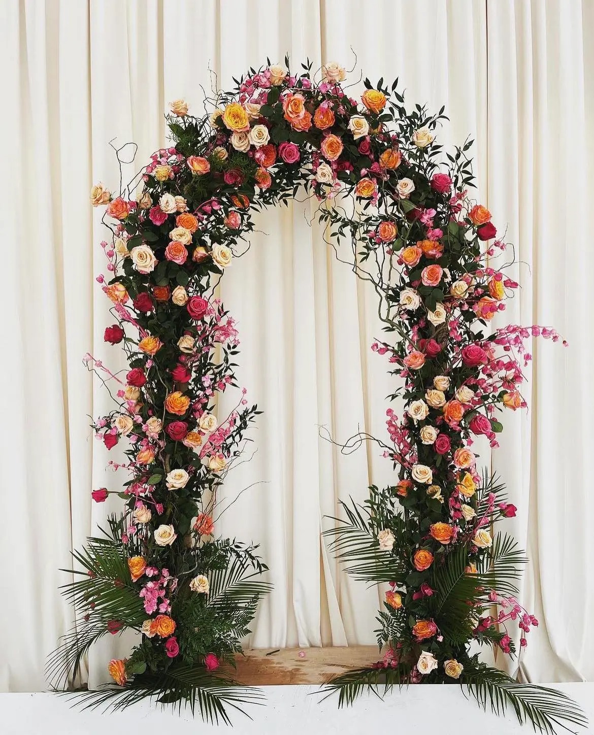 Vibrant wedding ceremony arch adorned with coral, pink, and yellow roses accented by dark foliage and tropical palm fronds