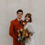 Bride and groom smiling against neutral wall, groom in rust-colored suit, bride holding orange and yellow bouquet