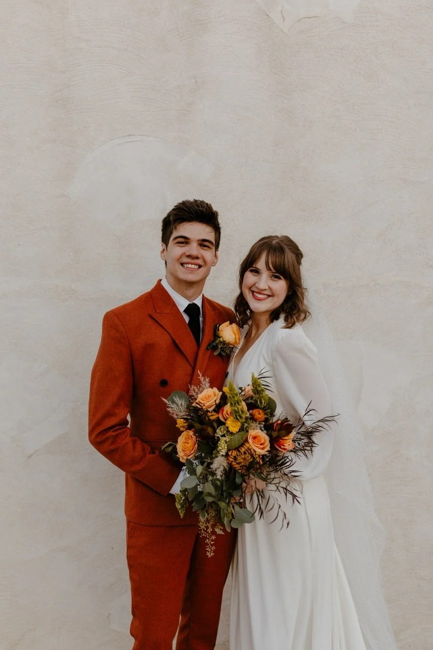 Bride and groom smiling against neutral wall, groom in rust-colored suit, bride holding orange and yellow bouquet