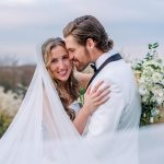 Bride with flowing veil and loose curled hair embracing groom in white tuxedo, holding white bouquet