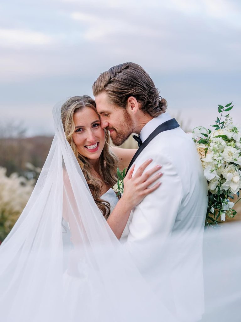 Bride with flowing veil and loose curled hair embracing groom in white tuxedo, holding white bouquet