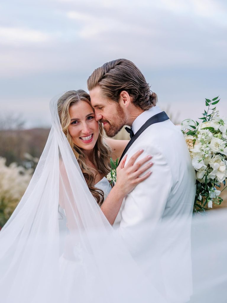 Bride with flowing veil and loose curled hair embracing groom in white tuxedo, holding white bouquet