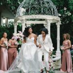 Two brides in white gown and suit walking down aisle from gazebo ceremony with pink-clad bridesmaids
