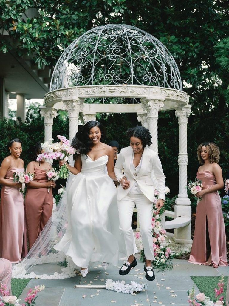 Two brides in white gown and suit walking down aisle from gazebo ceremony with pink-clad bridesmaids