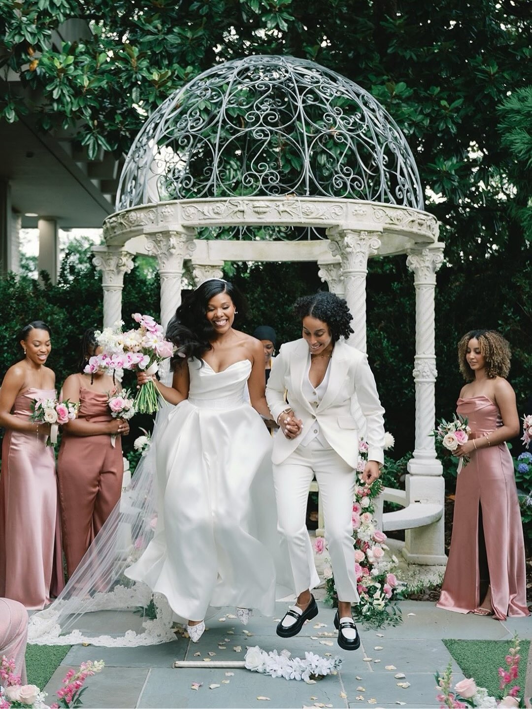Two brides in white gown and suit walking down aisle from gazebo ceremony with pink-clad bridesmaids