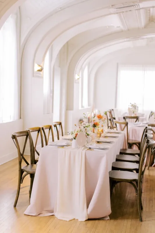 Elegant wedding reception table with blush linens, wooden cross-back chairs, and coral and white floral centerpieces in bright venue with arched ceilings