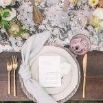 Overhead view of wedding place setting with menu card, pale blue napkin, gold flatware, and floral-patterned table runner