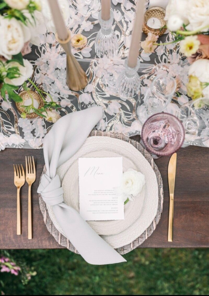 Overhead view of wedding place setting with menu card, pale blue napkin, gold flatware, and floral-patterned table runner