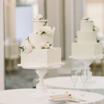 Three-tier square white wedding cake with blush roses and greenery on white cake stand