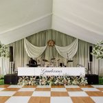 Wedding reception band stage with Gunderson's sign, floral garland, and checkerboard dance floor in tent