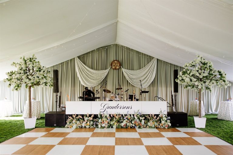 Wedding reception band stage with Gunderson's sign, floral garland, and checkerboard dance floor in tent