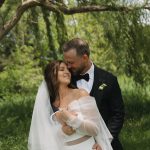 Groom carrying bride beneath weeping willow trees in romantic outdoor setting