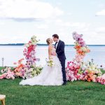 Bride and groom kissing at waterfront ceremony with elaborate colorful floral arch and ground arrangements