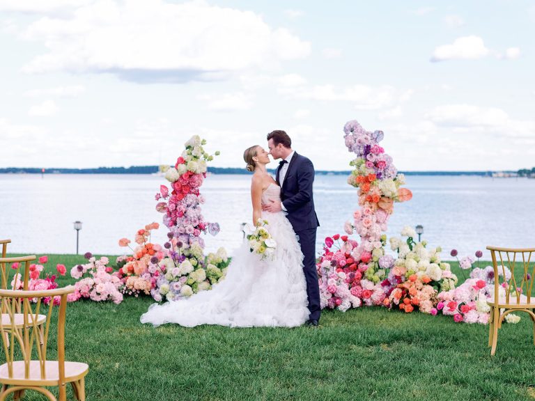 Bride and groom kissing at waterfront ceremony with elaborate colorful floral arch and ground arrangements