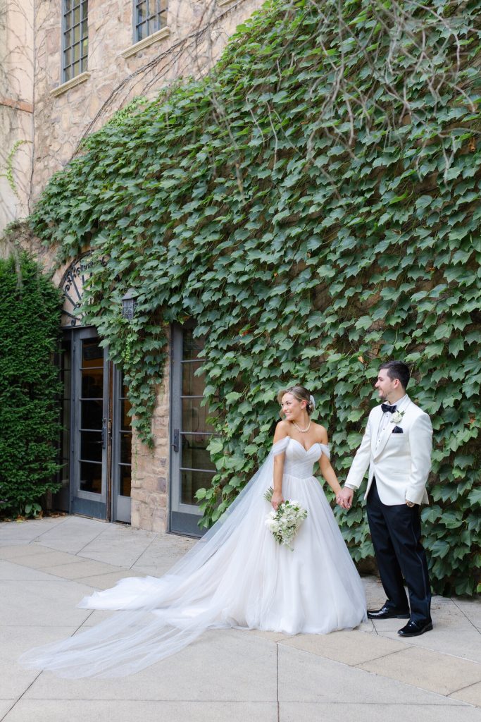 Bride in ballgown wedding dress and groom in white dinner jacket walking hand-in-hand before ivy-covered stone building