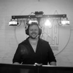 Smiling DJ in headphones behind turntables with stage lights and logo backdrop