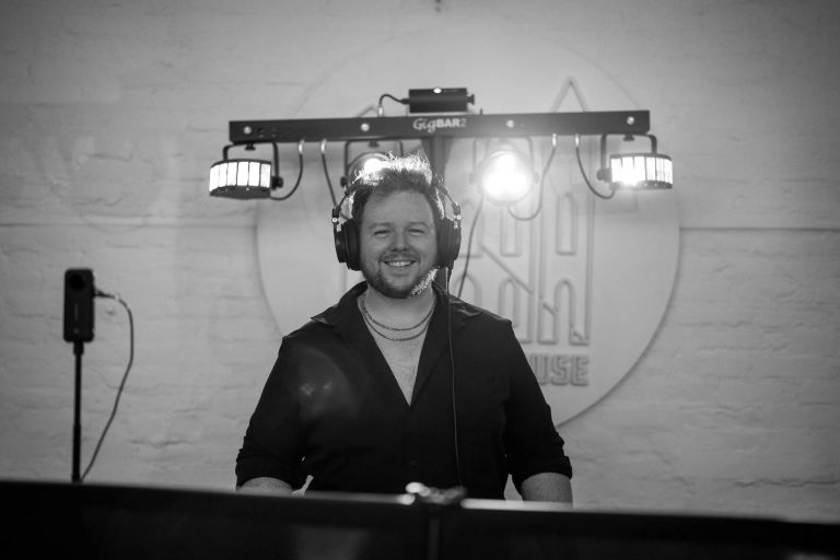 Smiling DJ in headphones behind turntables with stage lights and logo backdrop