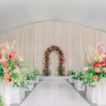 Indoor wedding ceremony aisle with floral arch, draped fabric backdrop, and colorful floral arrangements