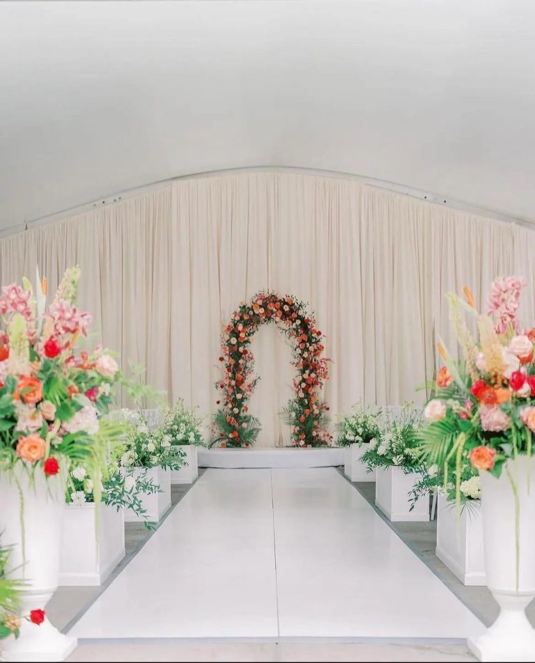 Indoor wedding ceremony aisle with floral arch, draped fabric backdrop, and colorful floral arrangements