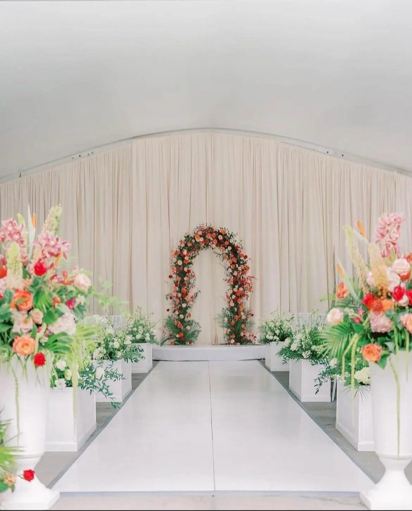 Indoor wedding ceremony aisle with floral arch, draped fabric backdrop, and colorful floral arrangements
