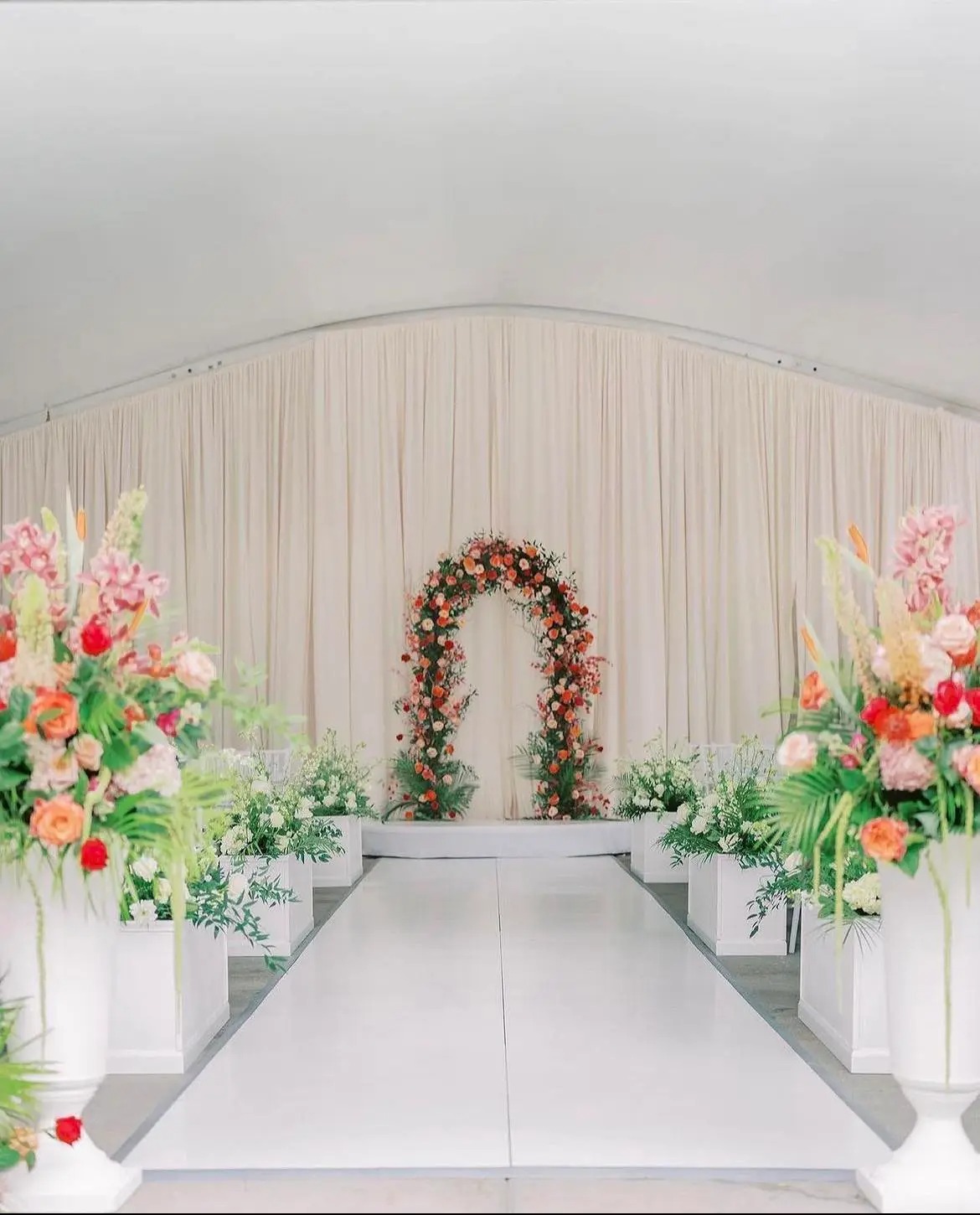 Indoor wedding ceremony aisle with floral arch, draped fabric backdrop, and colorful floral arrangements
