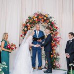Wedding ceremony with couple under vibrant circular floral arch featuring orange, red, and pink blooms against white drapery backdrop