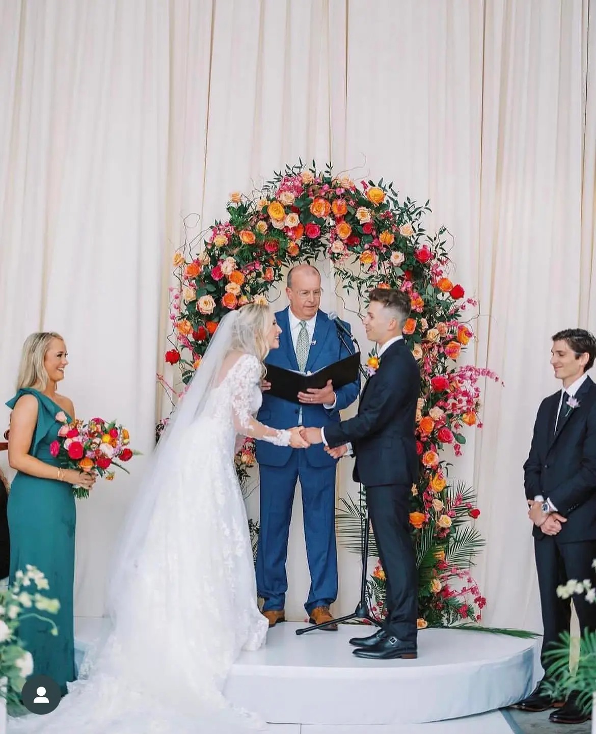 Wedding ceremony with couple under vibrant circular floral arch featuring orange, red, and pink blooms against white drapery backdrop