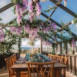 Outdoor dining table under pergola draped with purple wisteria and greenery overlooking waterfront