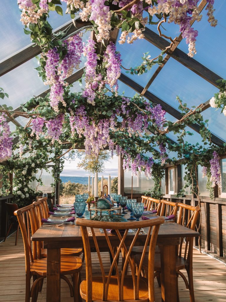 Outdoor dining table under pergola draped with purple wisteria and greenery overlooking waterfront