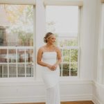 Smiling bride in white gown looking down near windows in light-filled room with wooden floor