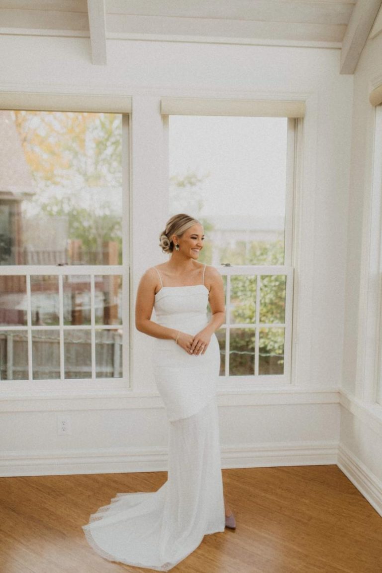 Smiling bride in white gown looking down near windows in light-filled room with wooden floor
