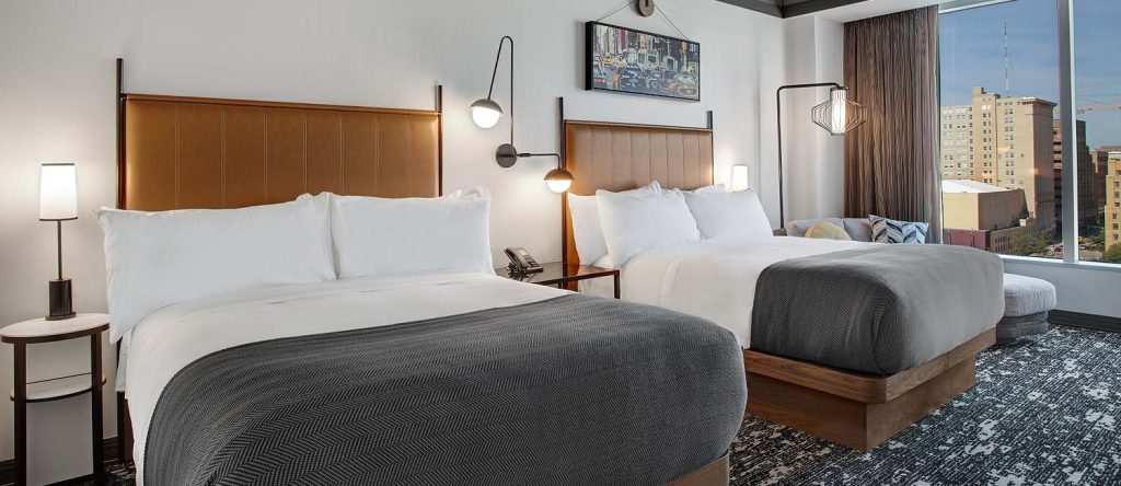 Modern hotel room with two queen beds, wooden headboards, gray accent throws, and city views