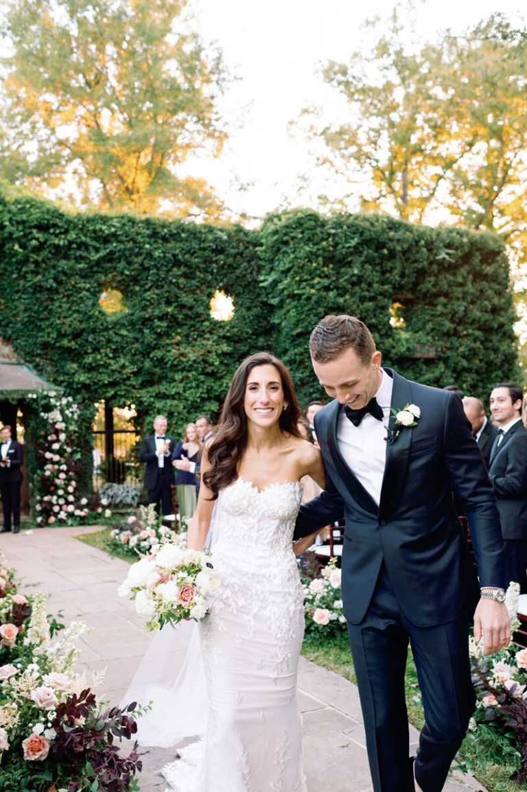 Newlyweds walking down aisle after ceremony in garden venue with floral-lined pathway and ivy-covered hedge backdrop